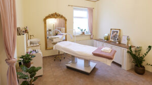 Behandlungsraum mit Liege, großem goldenen Spiegel, weißen Möbeln und sanfter, eleganter Einrichtung.