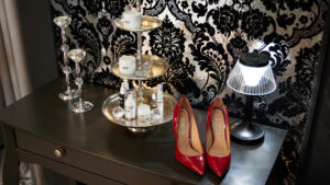 Dekoratives Arrangement mit Kosmetikprodukten, Kristallelementen und roten Pumps vor einer glänzenden, gemusterten Wand.